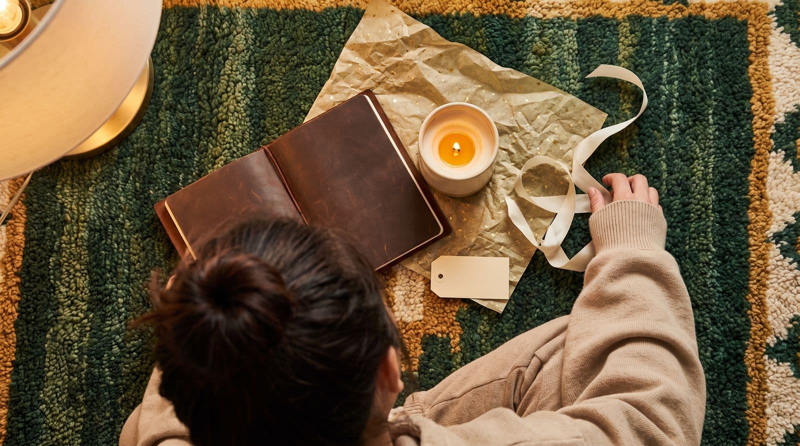 A young East Asian woman in an oversized hoodie reaches for a beautifully wrapped leather journal and a scented candle on a textured rug in a warm, cozy living room.