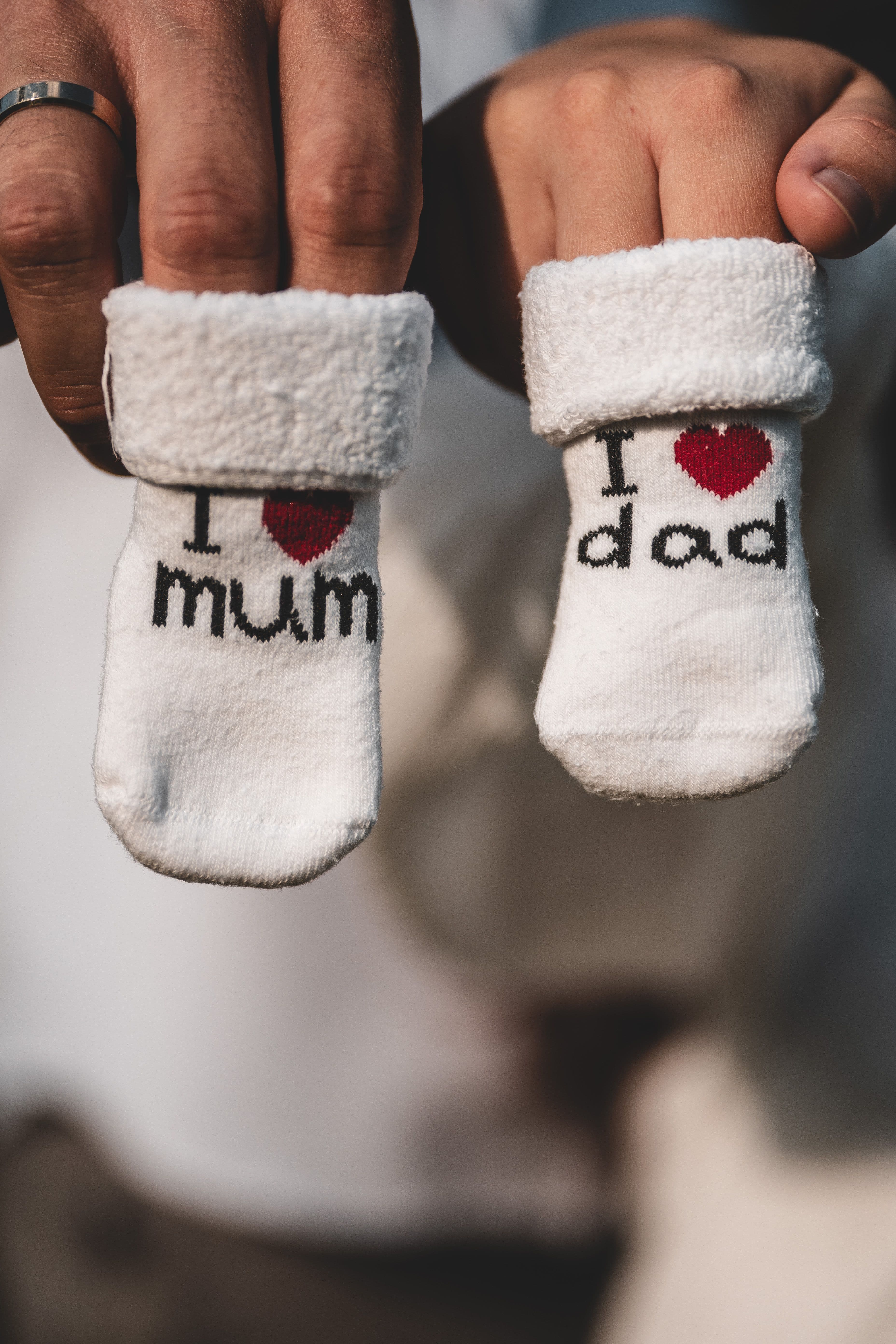 A person's hands holding up two white baby socks printed with 'I love mum' and 'I love dad,' each with a red heart design.