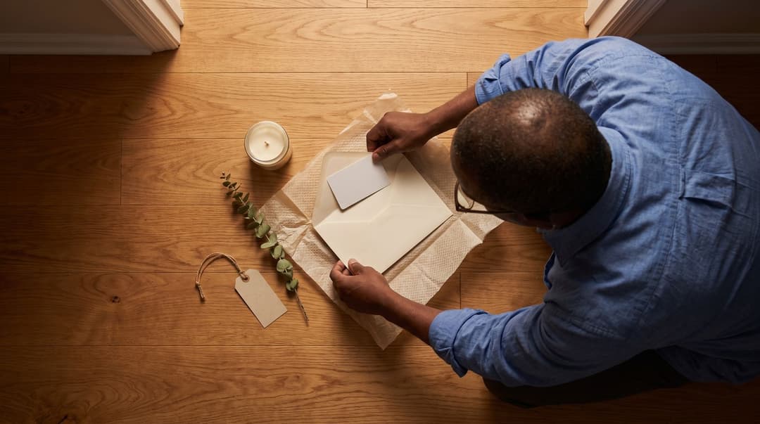 A man kneels on his new home's hardwood floor, opening a gift containing a local museum membership to help him settle into his new city.