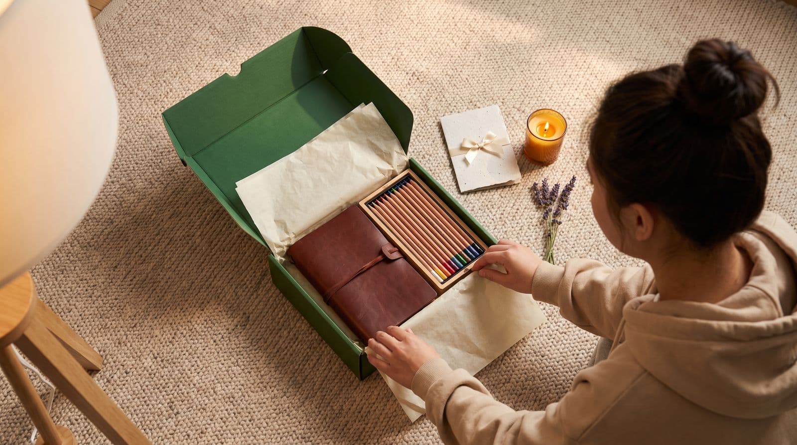 A woman in a cozy hoodie sits on a plush rug opening a thoughtful sabbatical gift consisting of a leather-bound travel journal and art supplies.