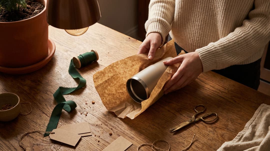 A woman's hands carefully wrap a high-quality insulated tumbler in paper on a wooden kitchen table, surrounded by gift-wrapping supplies.