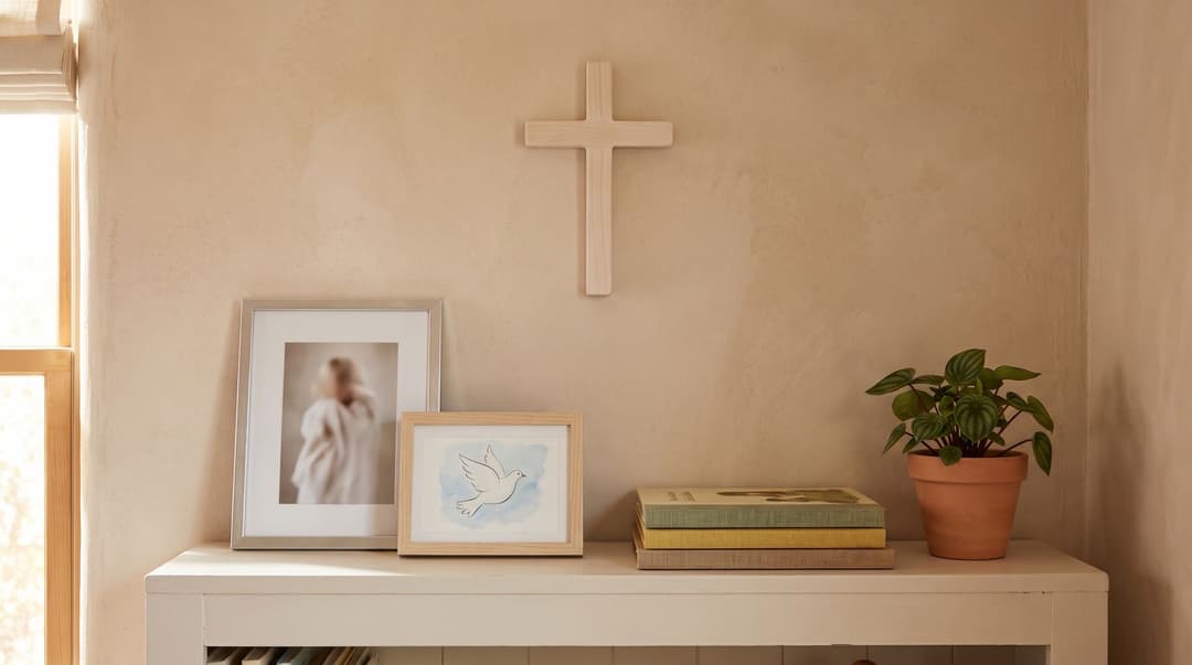 A pale wooden wall cross above a white shelf holding a silver frame with a soft, unrecognisable photo of a child in white, a small framed watercolour of a dove, a stack of books and a potted plant.