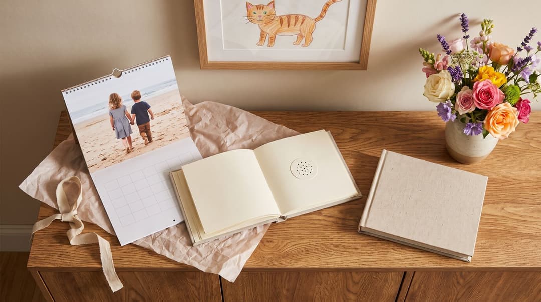 A pine sideboard under a framed crayon drawing of a cat, holding a wall calendar whose photo shows two children from behind on a beach, an open recordable storybook, a small speaker, a closed linen photo book and a jug of garden flowers.