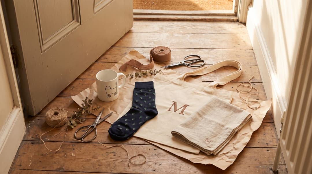 Gifts laid out on kraft paper on a sunlit wooden floor by an open door: a white mug with a line-drawn cat, navy socks with a tiny repeated plant motif, a natural canvas tote with a single M, a linen tea towel embroidered with a small M, ribbon, twine, scissors and a sprig of eucalyptus.
