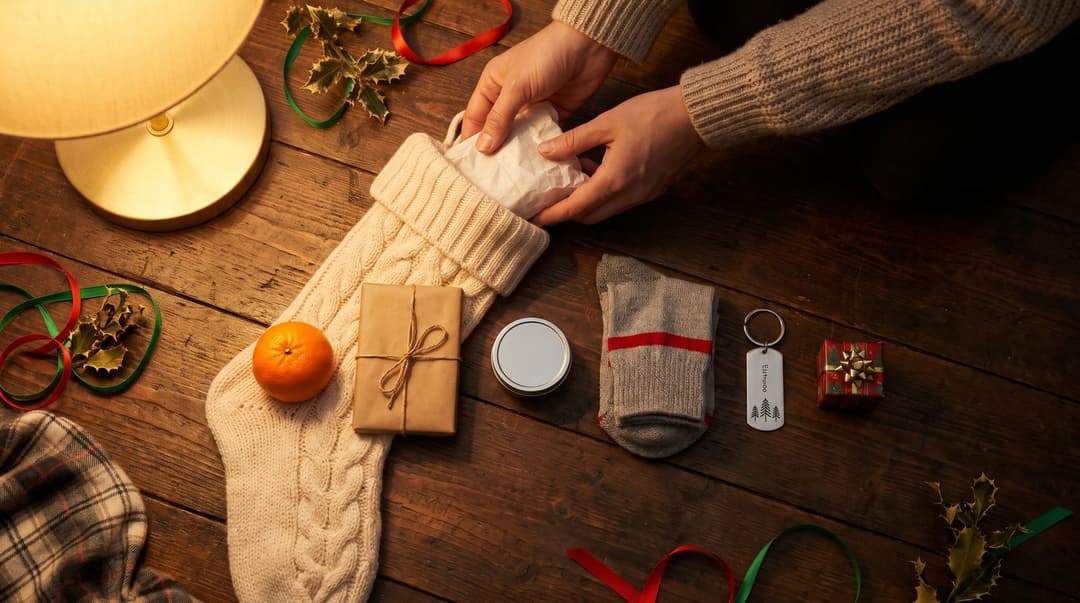 Hands tucking a tissue-wrapped parcel into a cream cable-knit stocking on a wooden floor in lamp light, with a clementine, a twine-tied parcel, a small tin, folded socks, an engraved keychain and a tiny wrapped box lined up beside it among holly and ribbon.