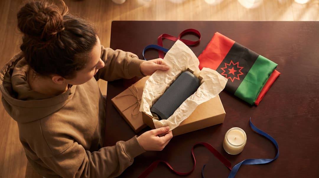 A woman in an oversized hoodie unwraps a gift box containing a portable speaker, with a Juneteenth flag and festive ribbons nearby on a wooden table.